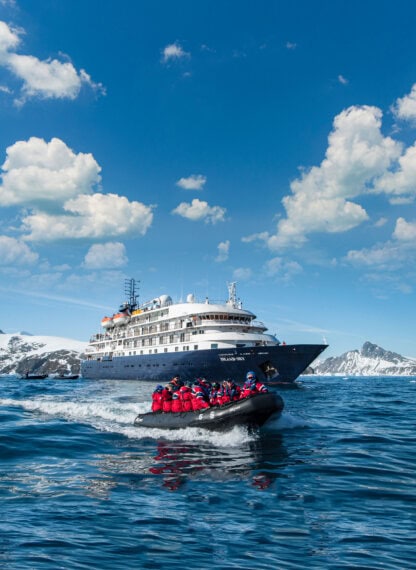 A group of people in red jackets ride a small inflatable boat on the ocean near a large cruise ship, embarking on an unforgettable Antarctica tour with snow-covered mountains in the background.