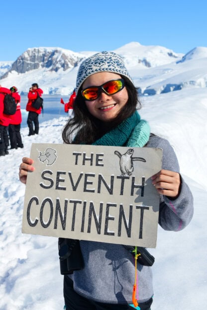 A woman in winter clothes and sunglasses holds a sign reading "The Seventh Continent" with a penguin illustration, standing on snow during an Antarctica tour, with mountains and people in the background.