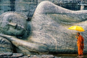 A monk in orange robes holds a yellow umbrella while standing in front of a large stone reclining Buddha statue—a serene moment often witnessed on a Sri Lanka cultural tour.
