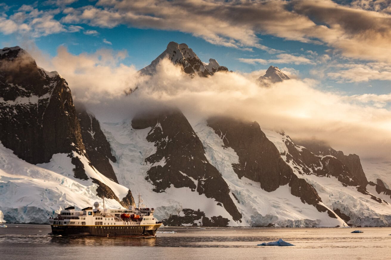 A cruise ship sails near snow-covered, rocky mountains with drifting clouds and icebergs in the water under a partly cloudy sky on an unforgettable Antarctica tour.