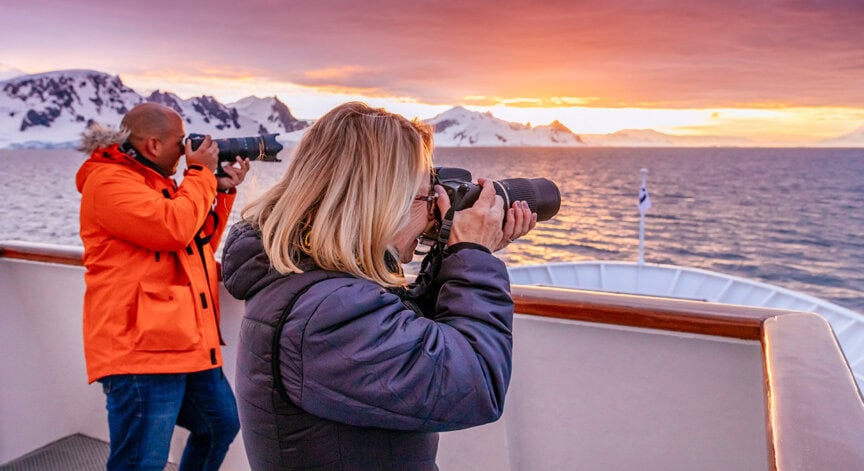 Two people on a ship’s deck photograph a snowy mountain landscape and colorful sky over the ocean at sunset during their Antarctica tour.